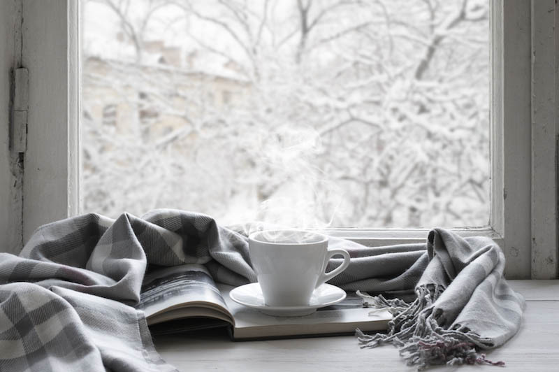 Cozy winter still life: cup of hot coffee and opened book with warm plaid on vintage windowsill against snow landscape from outside.