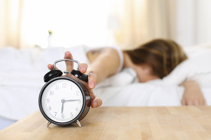 Sleepy young woman trying kill alarm clock while bury face in pillow. Early wake up, not getting enough sleep, getting work concept. Female stretching hand to ringing alarm willing turn it off